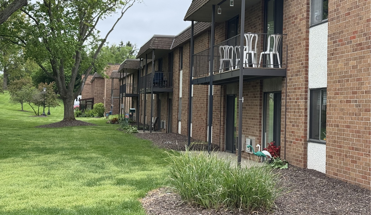 A view of an apartment building surrounded by a green lawn and trees, showcasing a welcoming outdoor space.